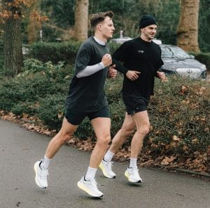 Två män springer tillsammans på en asfalterad väg i en park. De bär båda korta träningsskjorts och t-shirt över långärmad tröja. På fötterna har de vita och neongröna löparskor från Saucony. I bakgrunden syns bilar, gröna buskar och träd.