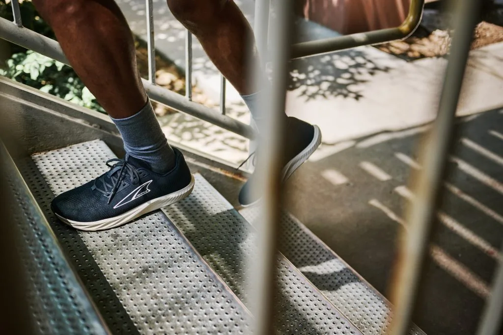 Person går uppför en metalltrappa i svarta löparskor med vit sula och grå strumpor. Bilden är tagen genom ett trappräcke i dagsljus.