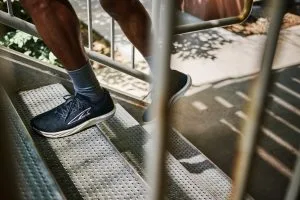 Person går uppför en metalltrappa i svarta löparskor med vit sula och grå strumpor. Bilden är tagen genom ett trappräcke i dagsljus.